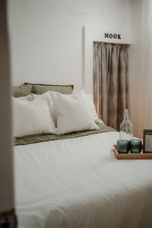 Een net opgemaakt bed met witte lakens en een groene deken, een nachtkastje met een fles en glazen, en een bordje boven het bed met de tekst 'NOOK'.