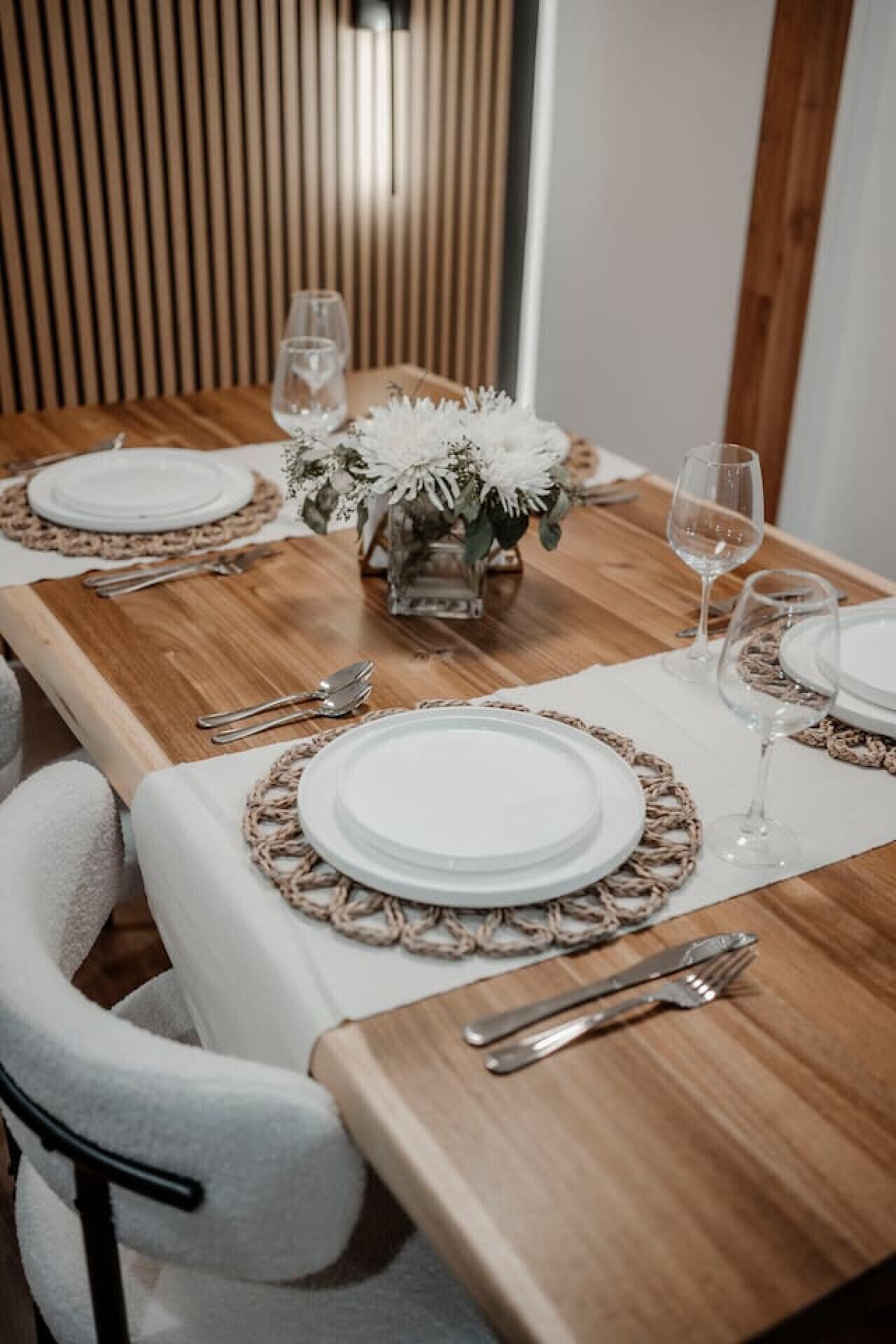 A dining table set with white plates, silverware, and wine glasses, centered with a floral arrangement.