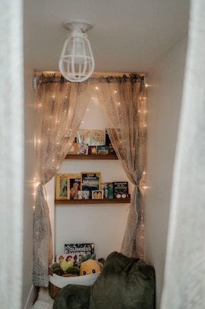 Un coin lecture confortable, agrémenté de rideaux à motifs, d’un luminaire au plafond et d’une bibliothèque remplie de divers livres et d’un jouet en peluche.