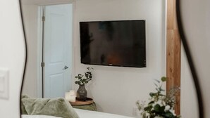 Een slaapkamer met een bed, een nachtkastje met een vaas, een aan de muur bevestigde televisie en een deur.