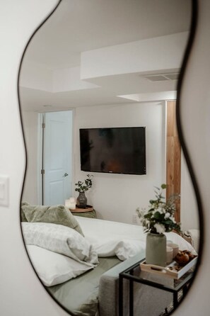 Une chambre à coucher avec un lit, une table de chevet ornée d’un vase, un téléviseur fixé au mur et une porte.