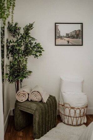 Una habitación con un reposapiés verde, una cesta de mimbre con cojines blancos, una manta beige enrollada y un cuadro en la pared.