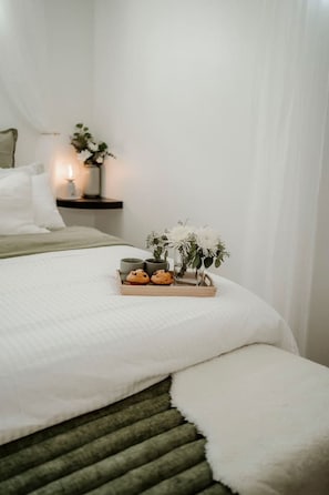 Een net opgemaakt bed met een witte dekbedovertrek, een houten dienblad met koekjes en een bloevas, en een nachtkastje met een kaars en een vaas.