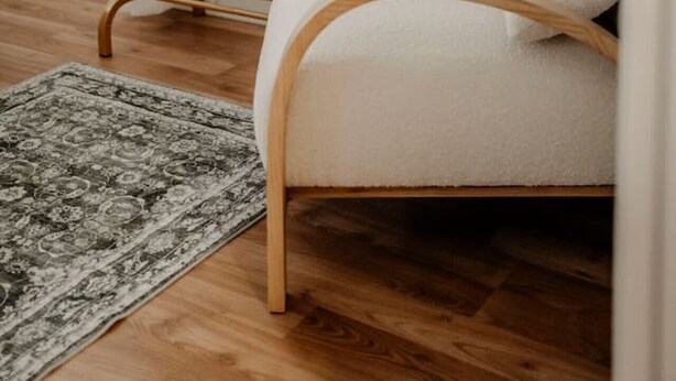 Een kamer met een houten vloer, een beige stoel, een houten stoel met een wit kussen, een beige badjas hangend aan een houten rek, en een kamerkleed met een patroon.
