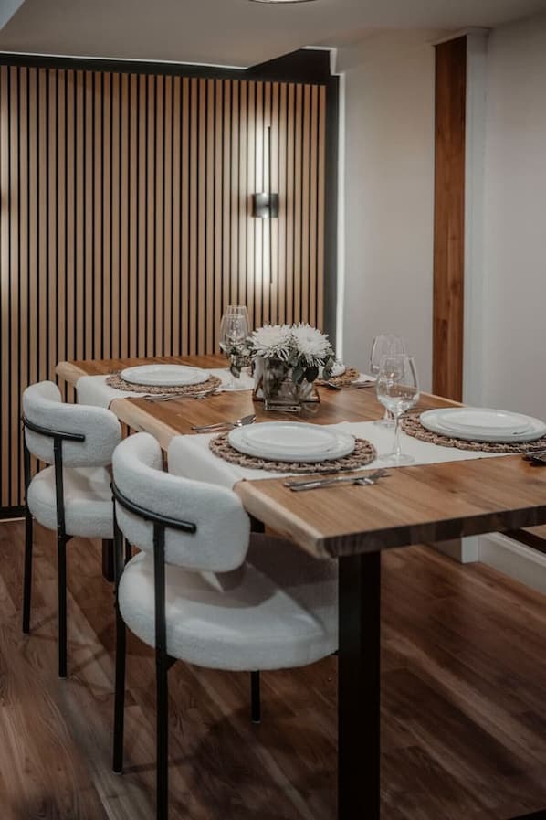 Comedor con mesa de madera dispuesta para seis personas, sillas blancas tapizadas y pared de listones de madera verticales.