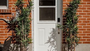 Een witte deur met een klein raam, aan weerszijden omgeven door twee planten in potten, en een deurmat met de tekst 'hello' op een betonnen pad.