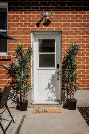 Une porte blanche avec une petite fenêtre, flanquée de deux plantes en pot, et un paillasson sur lequel est écrit le mot « bonjour », le tout placé sur un chemin de béton.