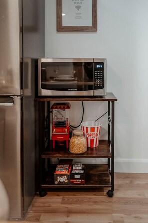 A kitchen with a stainless steel refrigerator, a microwave on a wooden shelf, a popcorn machine, a popcorn bucket, and snack boxes.