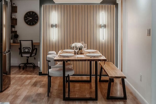 Een eethoek met een houten tafel voor vier personen, een klok aan de muur, en een moderne keuken op de achtergrond.