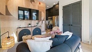 Un salon moderne avec un canapé, une table basse et un téléviseur fixé au mur. Il y a également un coin cuisine équipé d’un four à micro-ondes et d’un réfrigérateur.