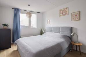 A bedroom with a bed, a bedside table with a lamp, a window with curtains, and two framed pictures on the wall.