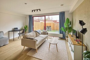 A modern living room with a sofa, a coffee table, a desk with a laptop, and large windows with curtains.