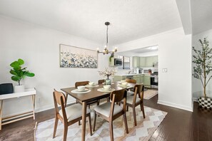 A dining area with a wooden table, chairs, and a chandelier. There is a plant on the left and a painting on the wall.