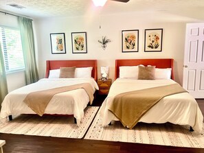 A hotel room with two beds, each with a red headboard, white bedding, and a beige throw blanket. There are three framed floral artworks on the wall, a small bedside table with a lamp, and a door on the right.