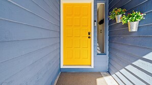 A yellow door with a white frame, a doormat, and two metal buckets with flowers on a blue siding wall.