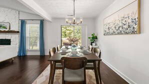 A dining room with a wooden table set for six, a chandelier, and a large window with blue curtains.