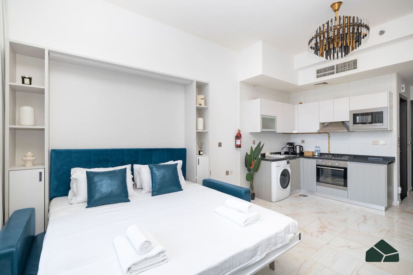 Una habitación de hotel moderna con una cama grande, cabecera azul, lavadora integrada, kitchenette y un candelabro.