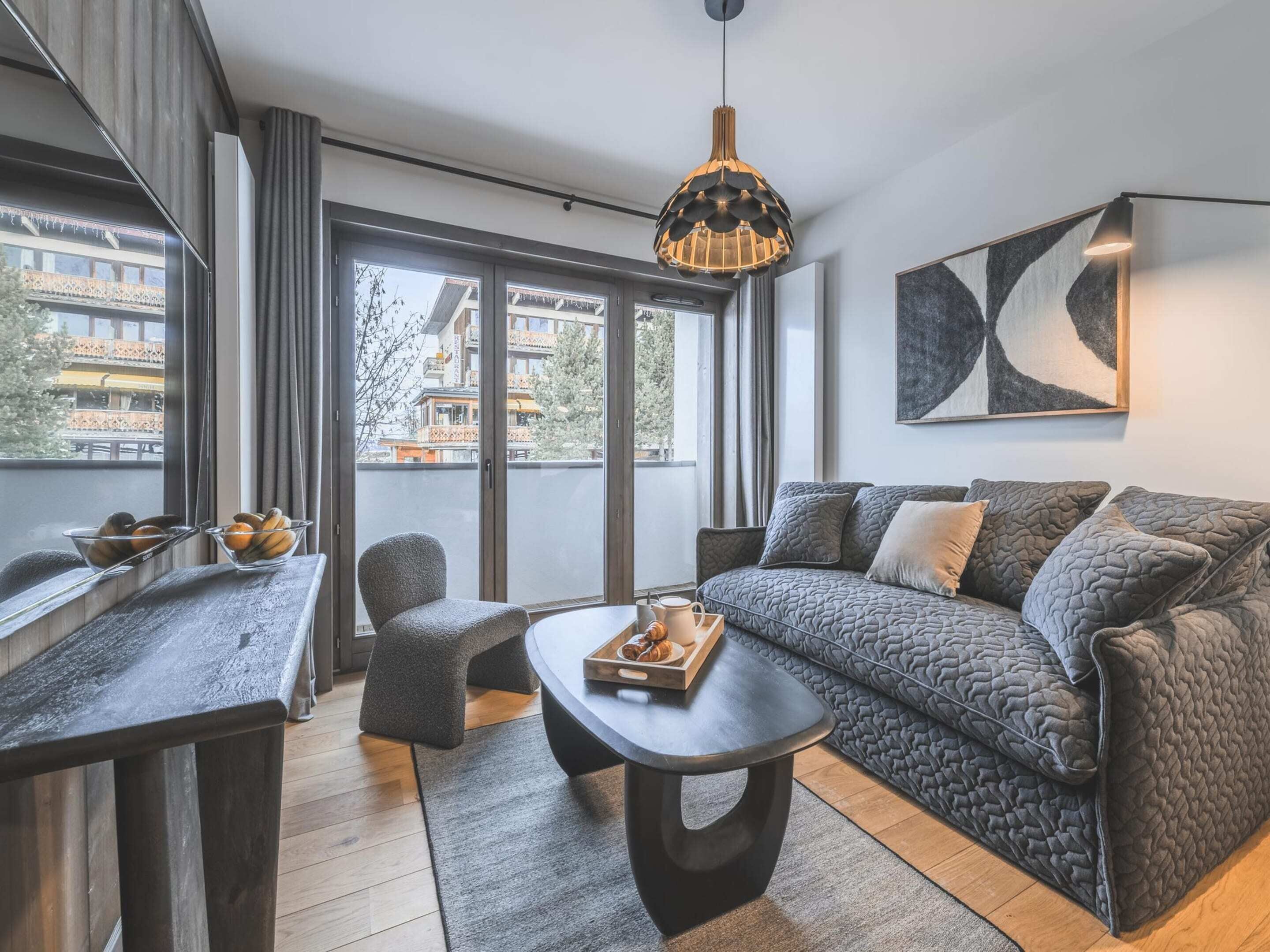 Un salon moderne comprenant un canapé, une table basse, un fauteuil et une table à manger. Une grande fenêtre offre une vue sur les bâtiments et les arbres.