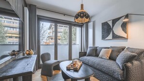 A modern living room with a sofa, a coffee table, a chair, and a dining table. There is a large window with a view of buildings and trees. - Apartment in Plan Peisey Near Vanoise Park (Peisey-Nancroix)