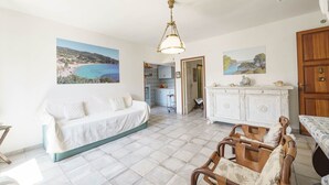 Ein großzügiges Wohnzimmer mit einem weißen Sofa, einem Esstisch mit Stühlen, einer weißen Kommode und einer Deckenleuchte. An den Wänden hängen zwei Gemälde, und eine Tür führt in einen anderen Raum.