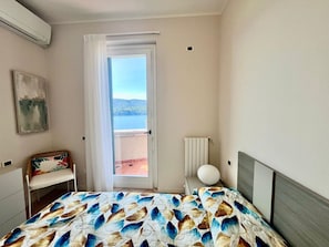 A bedroom with a bed, a chair, a radiator, and a view of the sea through a door.