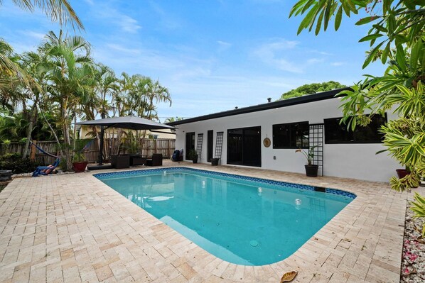 A swimming pool with a tiled deck, a covered seating area, and a modern building with large windows.