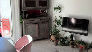 A living room with a TV mounted on the wall, a dining table with chairs, a cabinet with glass doors, and several potted plants. - Cosy village house (Peynier)
