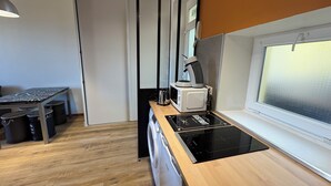 Kompakti keittiö, jossa on musta induktioliesi, valkoinen mikroaaltouuni ja kahvinkeitin työtasolla. Nurkassa on puinen pöytä tuoleineen ja roskakori.
