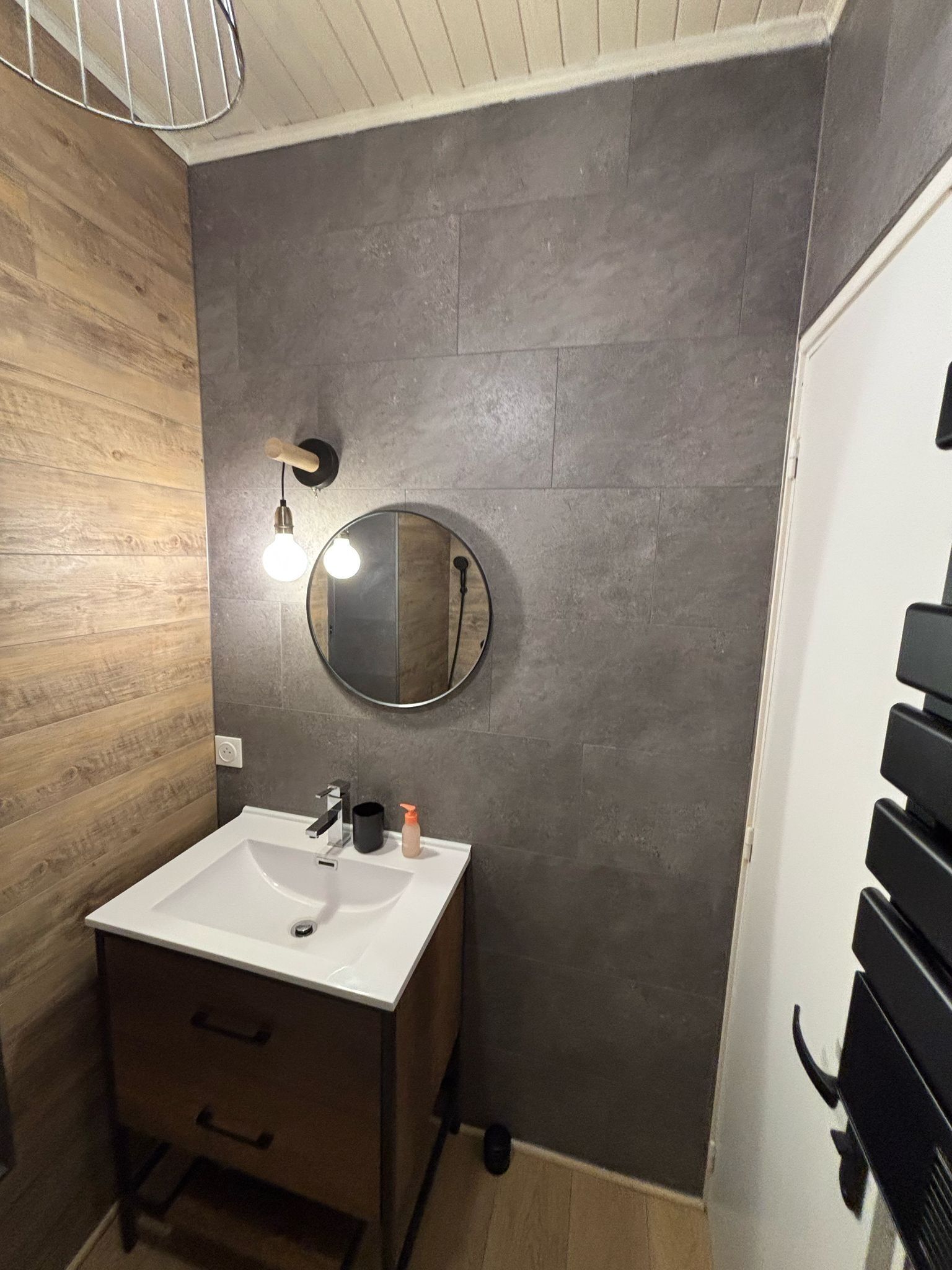 A modern bathroom with a wooden vanity, a round mirror, and a light fixture.