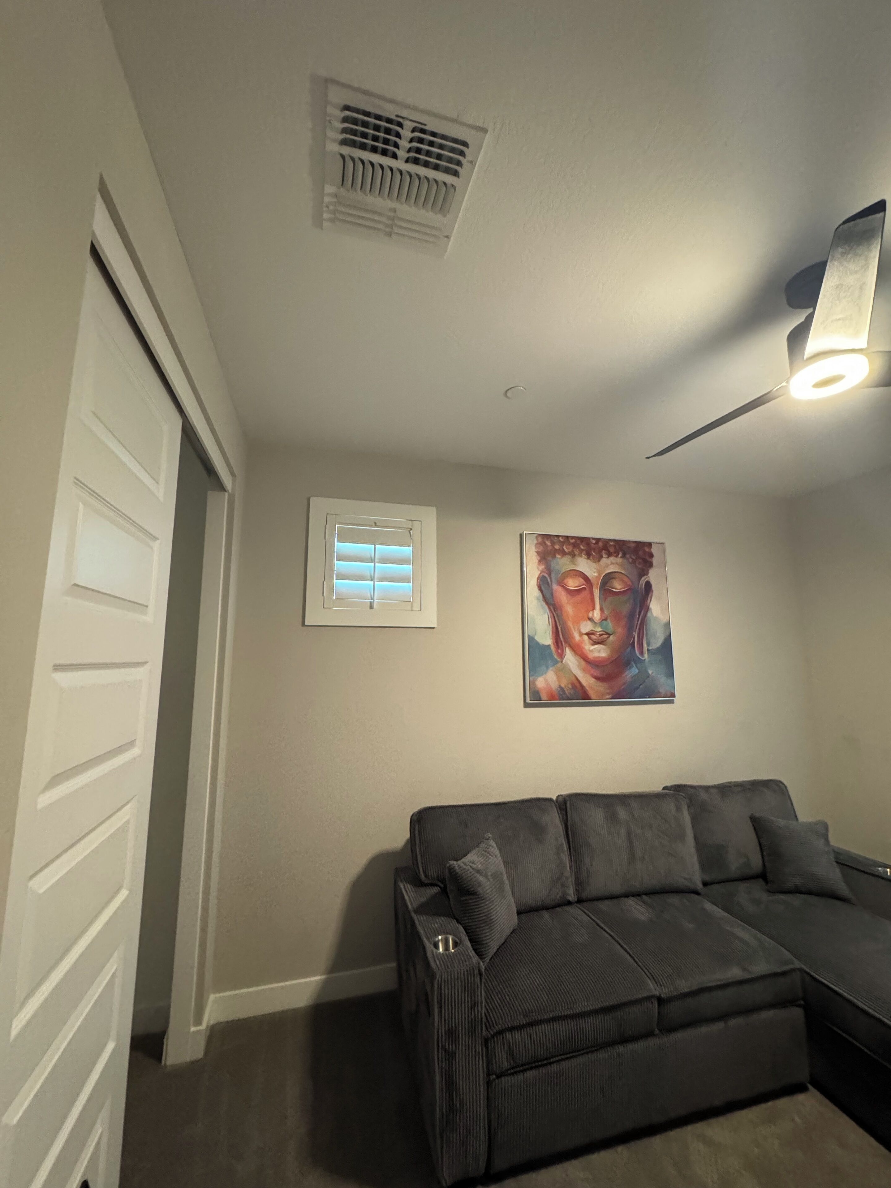 A room with a dark gray sectional sofa, a ceiling fan, a wall-mounted air vent, a window with blinds, and a Buddha painting.