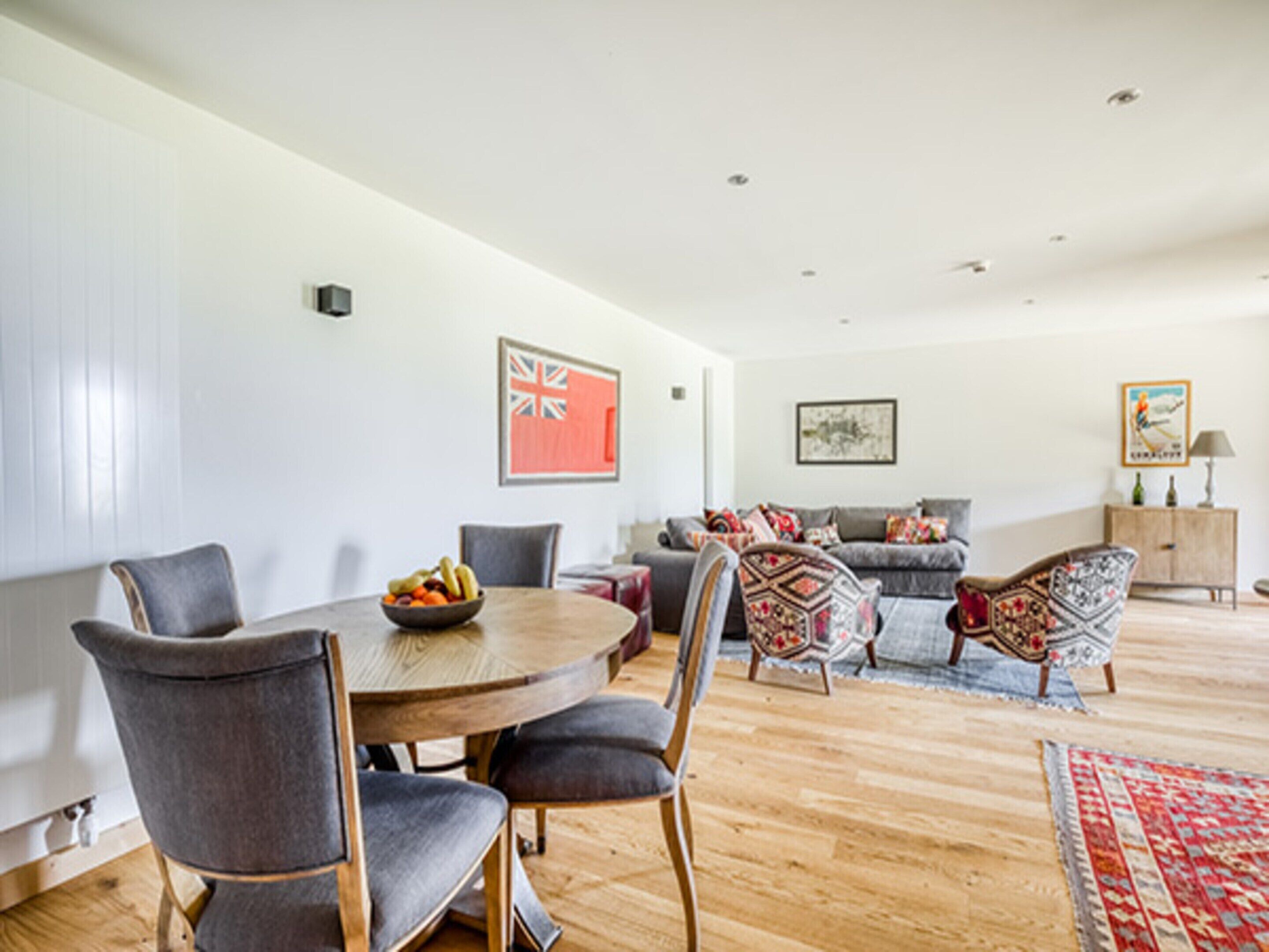 Un salon moderne avec un coin repas, comprenant une table ronde en bois, des chaises à revêtement gris, un ensemble de canapés gris et un tapis à motifs.