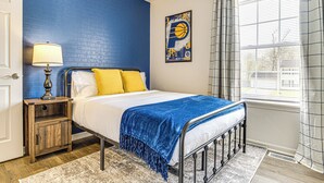 A bedroom with a bed, a nightstand, a lamp, a window with curtains, and a wall with a basketball-themed artwork. - 9 Mi to Dtwn Indianapolis: Family-Friendly Home! (Indianapolis)