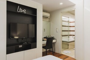 A modern hotel room with a flat-screen TV mounted on a black wall, a desk with a chair, a glass shower enclosure, and a white bathtub.