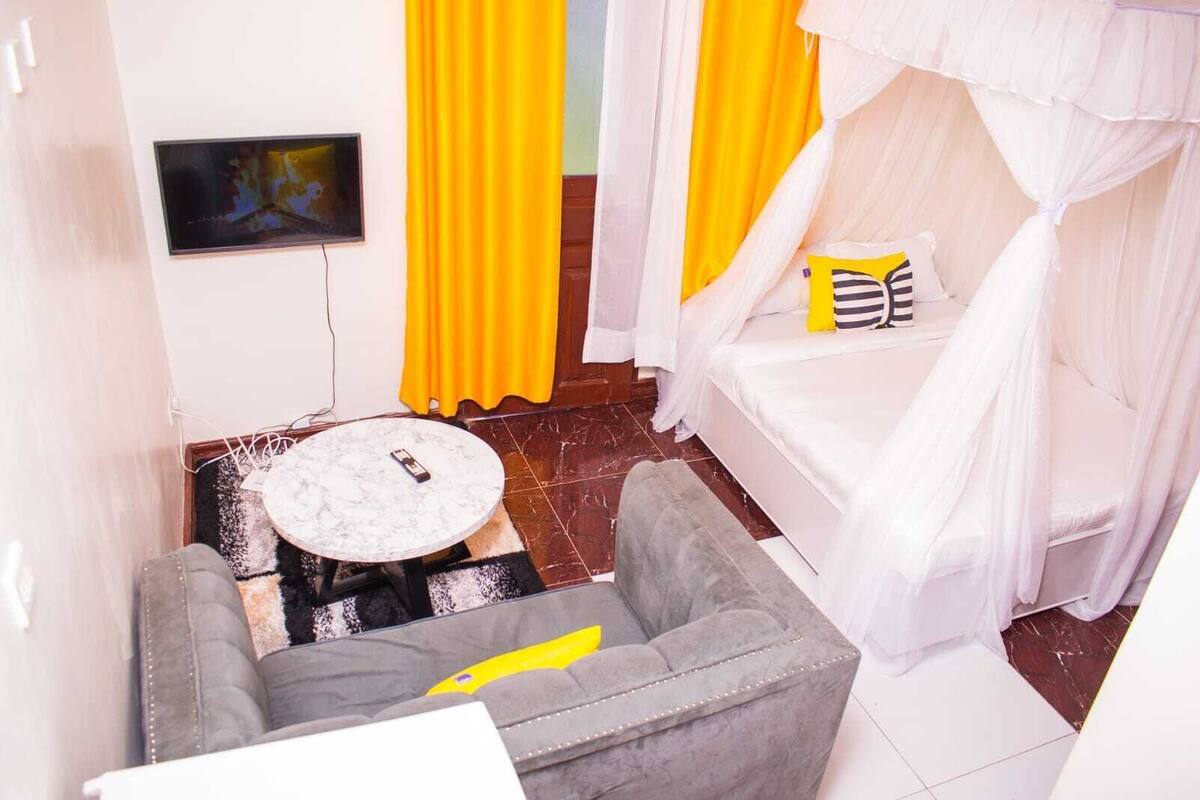 Habitación con cama cubierta por un dosel blanco, un sofá gris, una pequeña mesa redonda con un control remoto y cortinas amarillas.