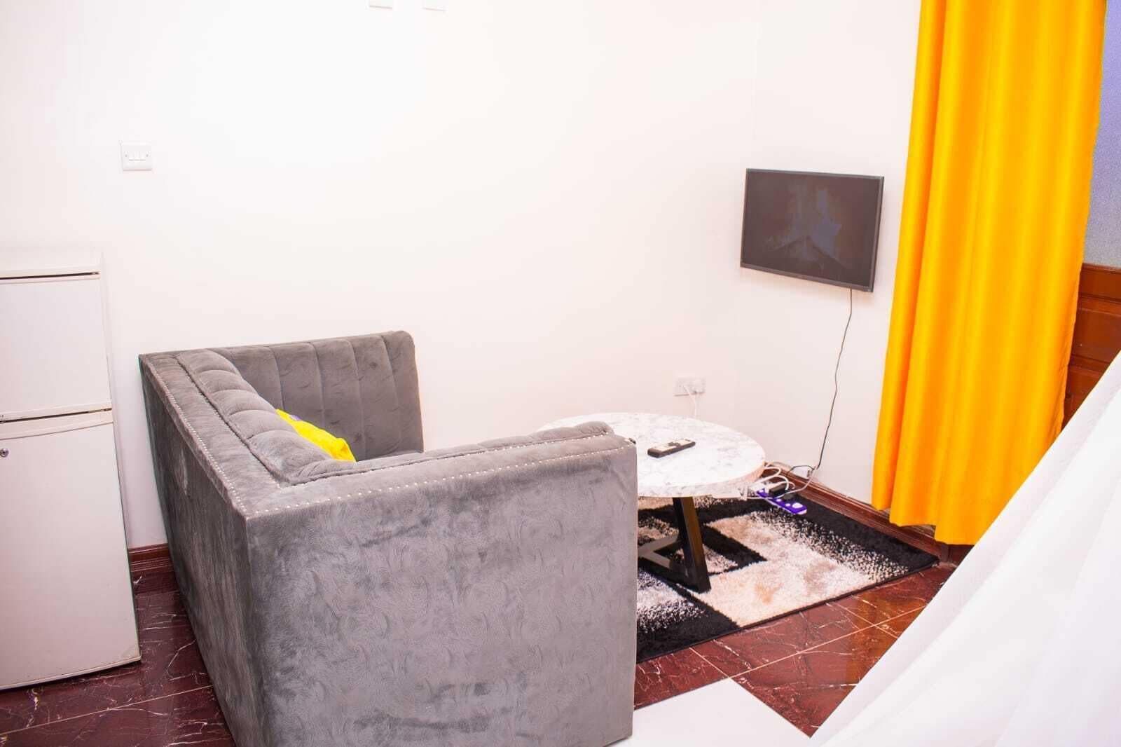 A small, compact room with a grey upholstered armchair, a small round table, and a television mounted on the wall.