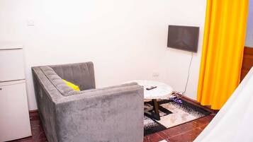 A small, compact room with a grey upholstered armchair, a small round table, and a television mounted on the wall.