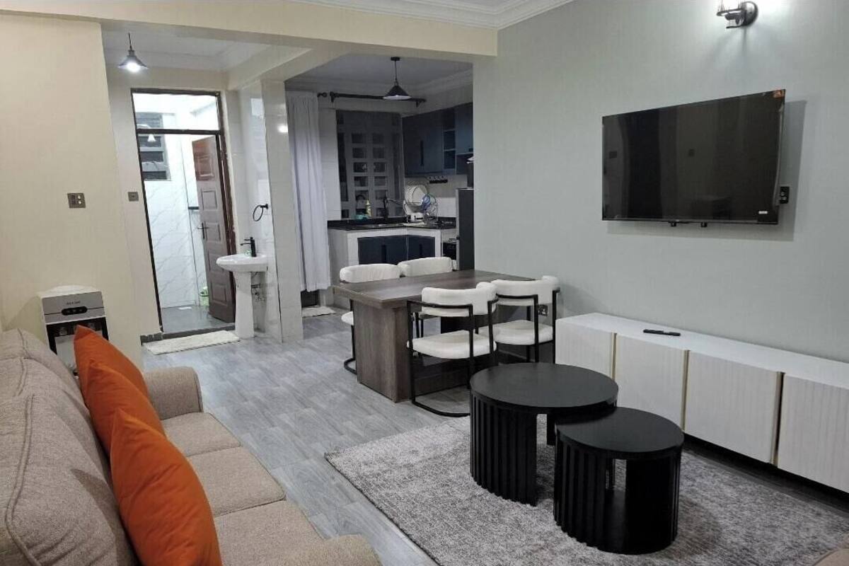 Una sala de estar moderna con un sofá, dos mesas auxiliares negras, una mesa de comedor con sillas, una zona de cocina y un televisor montado.