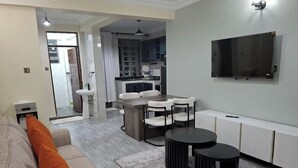 Ein modernes Wohnzimmer mit einer Couch, zwei schwarzen Beistelltischen, einem Esstisch mit Stühlen, einer Küchenzeile und einem Wand-Fernseher.