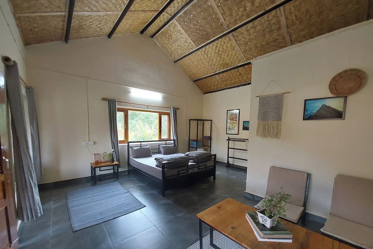 A room with a bed, a wooden table, a small desk, a window with curtains, a wall-mounted shelf, and a painting.
