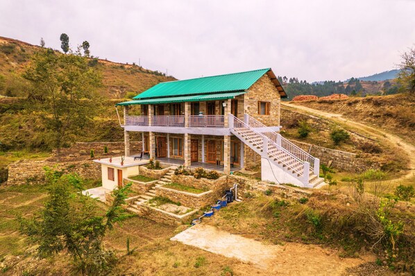 A two-story stone building with a green roof, a balcony, and a staircase leading to the upper floor. - Himkosi Farm by the River (Almora)