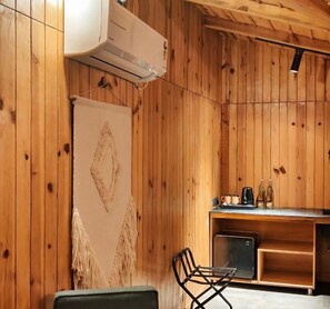 A wooden interior with a wall-mounted air conditioner, a hanging tapestry, a small kitchenette, and a seating area with a chair. - Himkosi Farm by the River (Almora)