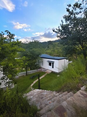 A small white building with a blue roof, surrounded by lush greenery and a white picket fence. - Himkosi Farm by the River (Almora)