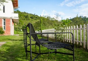 A metal bench with a slatted seat and backrest, positioned in a grassy area with a white picket fence in the background. - Himkosi Farm by the River (Almora)