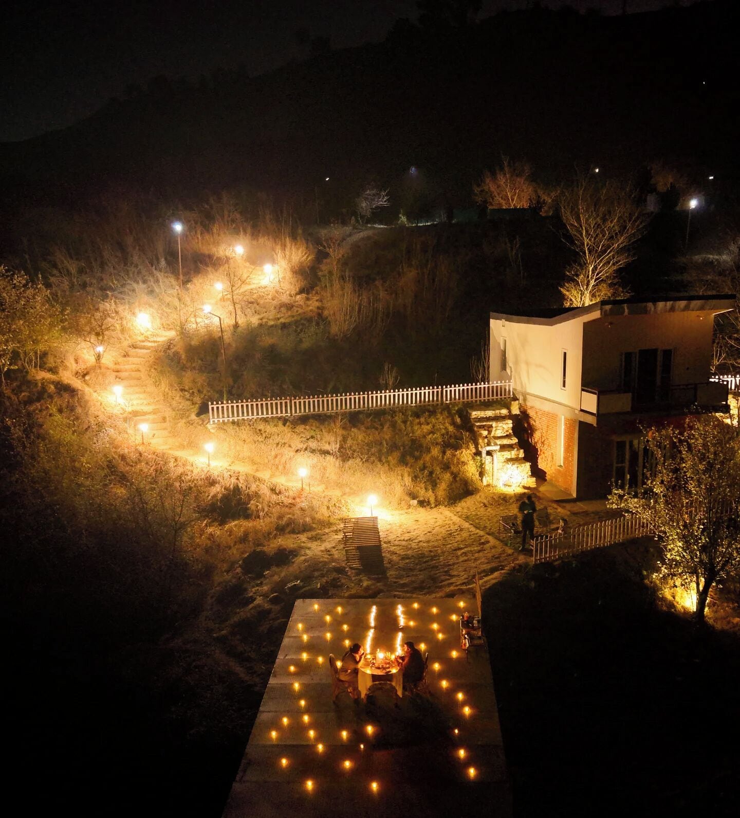 Ein Außenbereich bei Nacht mit einem gepflasterten Weg, einem kleinen Pool mit Kerzen und einem Gebäude mit weißem Zaun.