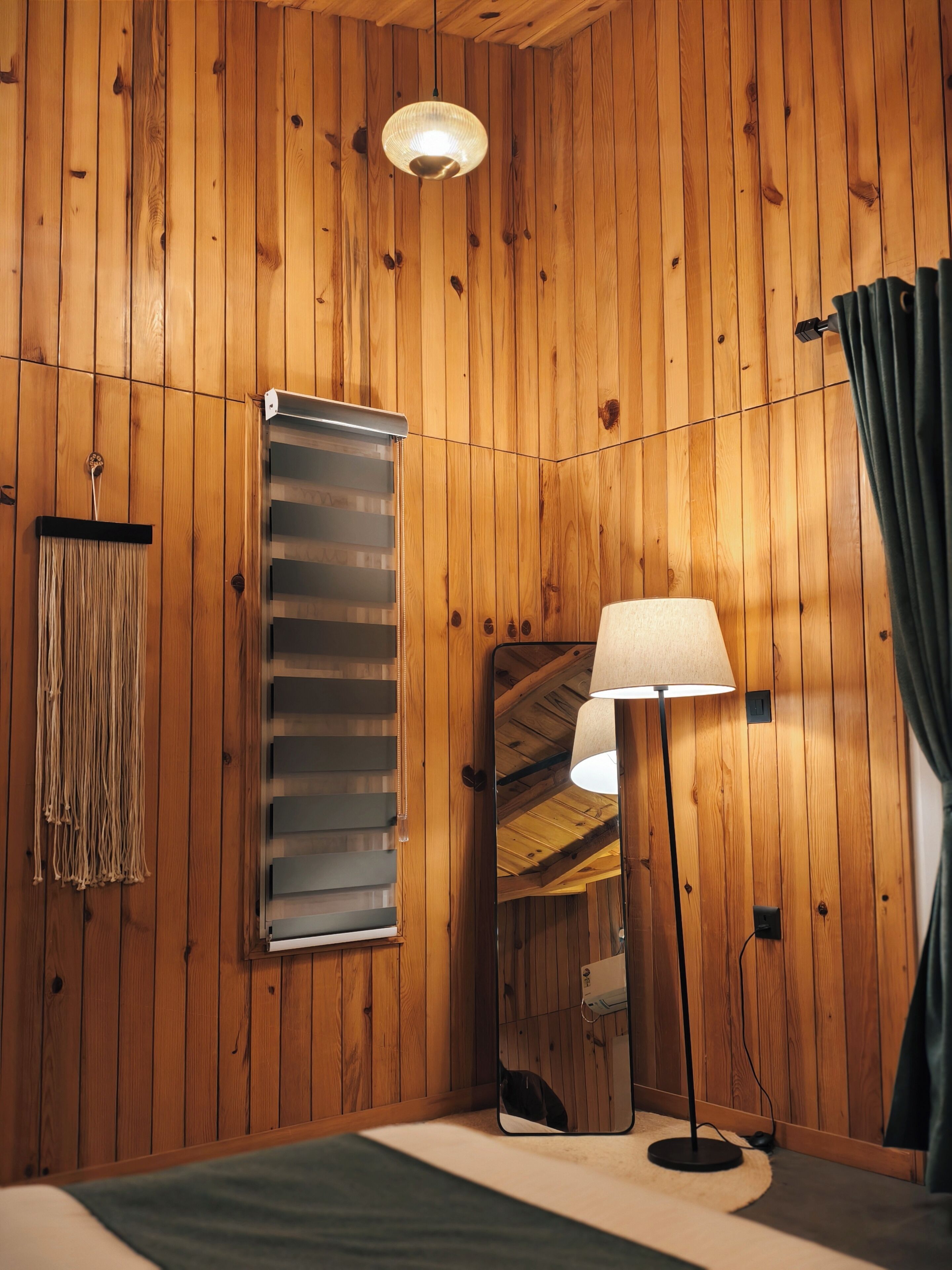 Ein Zimmer mit Holzwandverkleidung, einem Spiegel, einer Stehlampe, einem Fenster mit Jalousien und einem Vorhang.