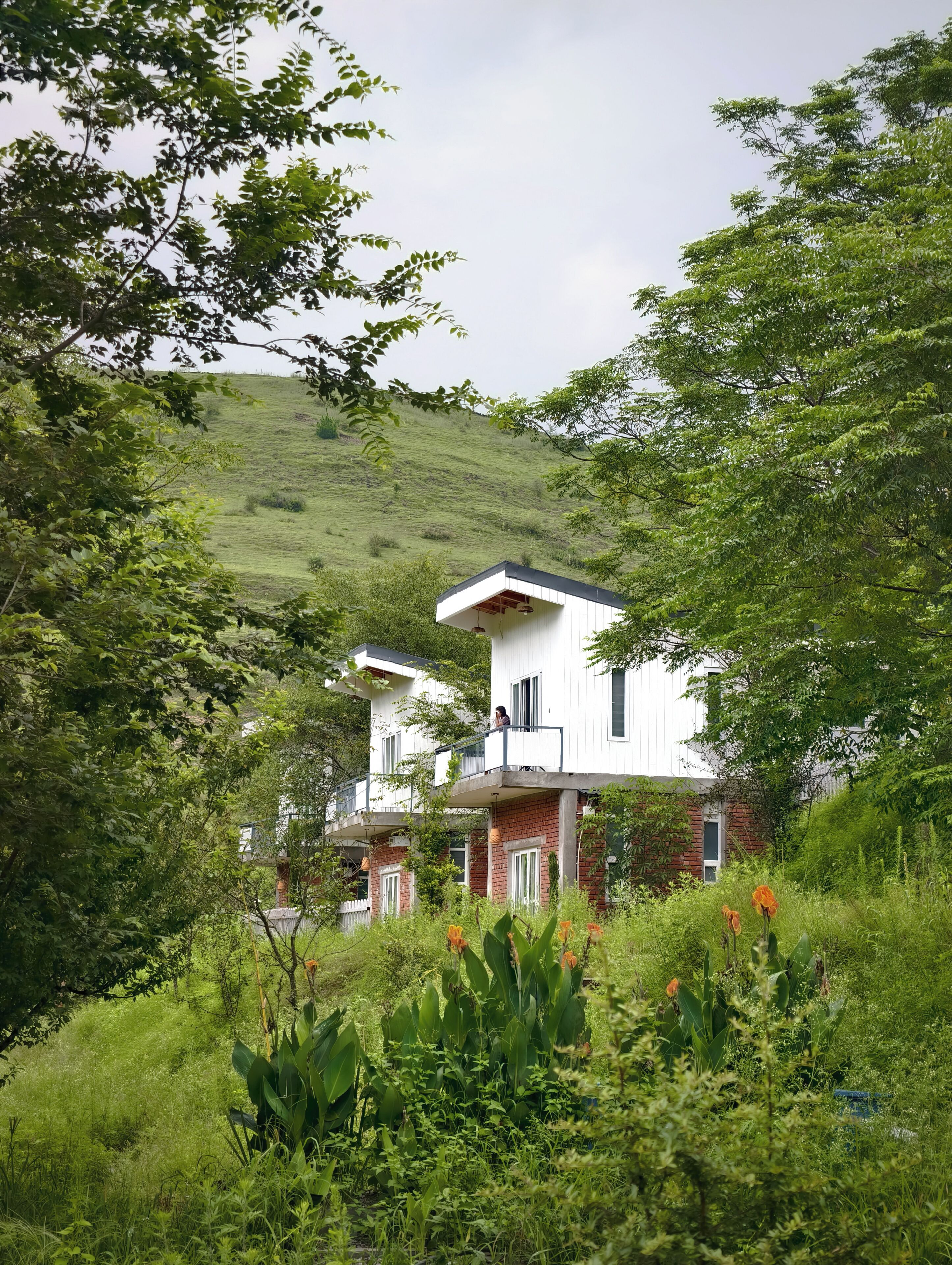 Un edificio de dos pisos con balcón, rodeado de exuberante vegetación y un paisaje montañoso.