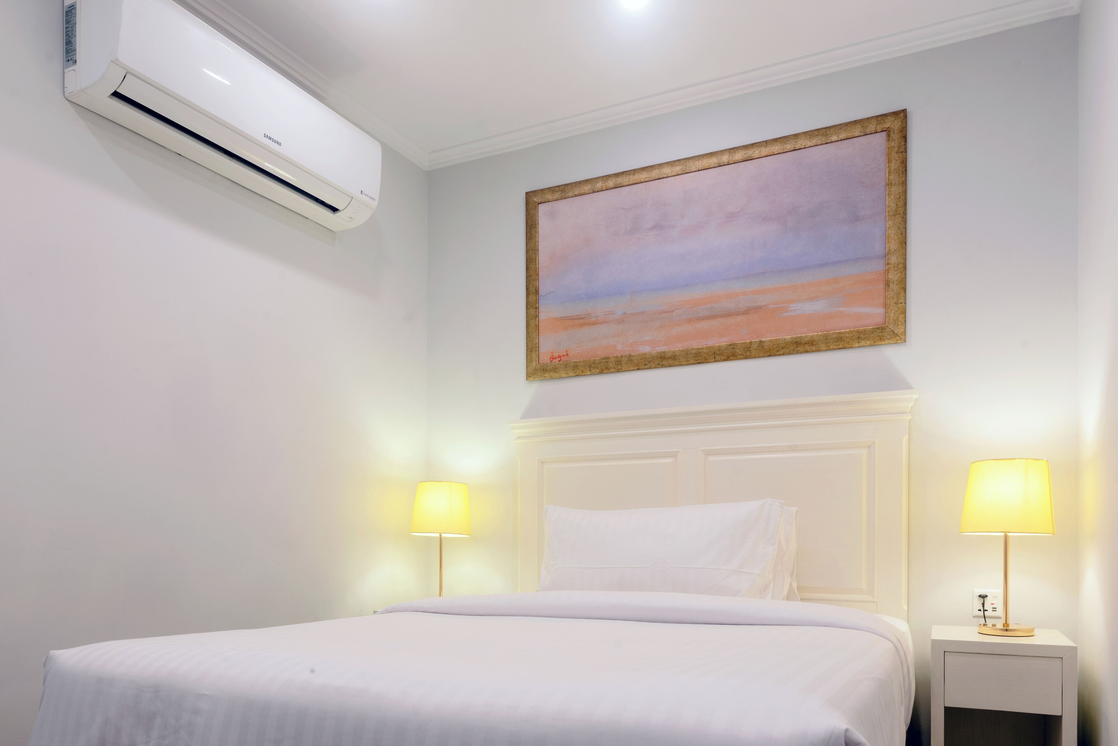 Une chambre d’hôtel avec un lit, deux lampes de chevet, un climatiseur et un tableau au mur.
