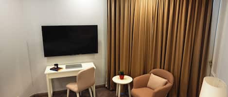 Habitación de hotel con televisor de pantalla plana, un escritorio con una computadora portátil, dos sillas, una mesita redonda con una planta y un ventanal con cortinas.