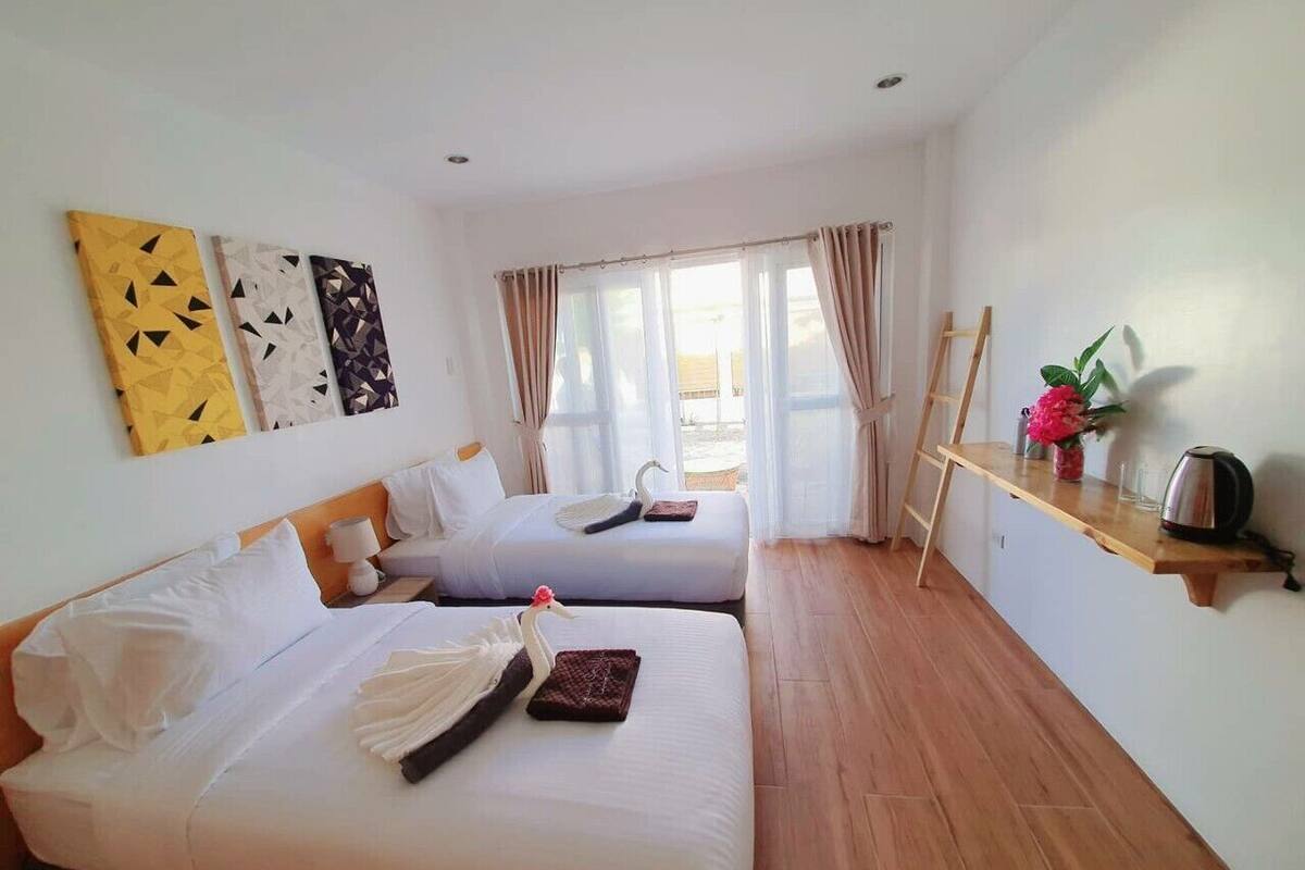 A hotel room with two beds, a wooden floor, a desk with a kettle, and a window with curtains.