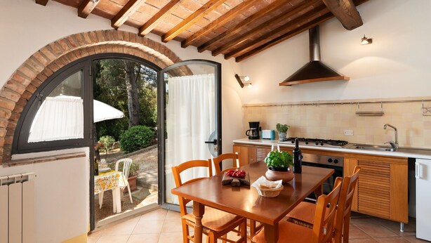 A cozy kitchen with a dining area, featuring a brick archway, wooden beams, and a view of the garden.